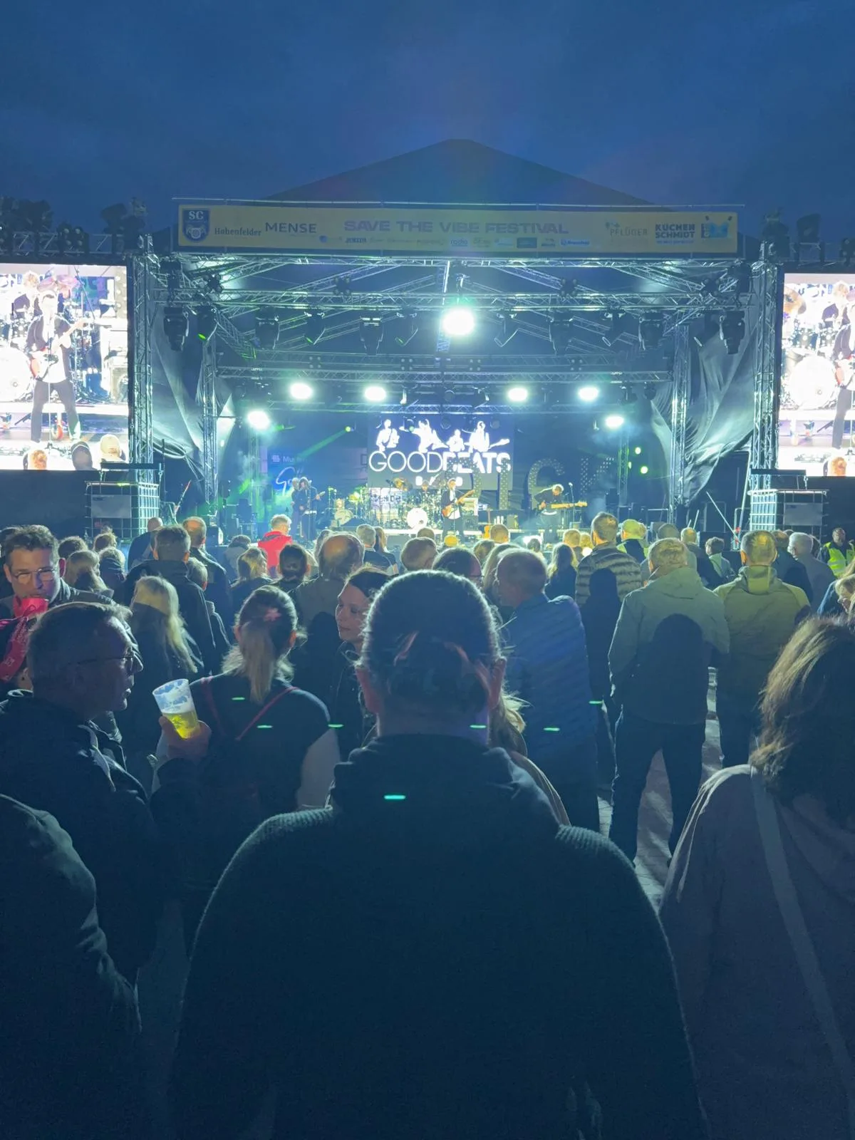 Blick aus dem Publikum auf die Open-Air-Bühne des Save-the-Vibe-Festivals am Abend, auf der Leinwand hinter der Band steht GOODBEATS.