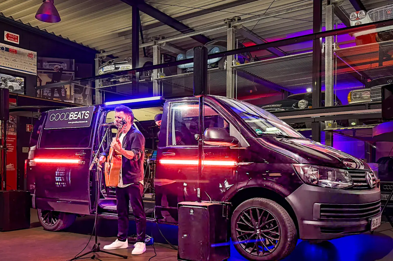 Das Konzert-Taxi in einer Halle mit Oldtimern: Vor der offenen Seitentür des GOODBEATS-Bullis singt ein Musiker mit Akustikgitarre, im Bulli sitzt ein zweiter.