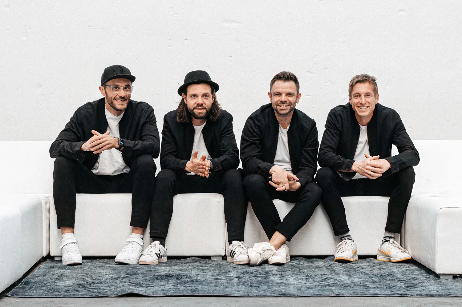 Die vier Musiker der GOODBEATS sitzen in schwarzen Jacken und weißen Shirts nebeneinander auf einem weißen Sofa.
